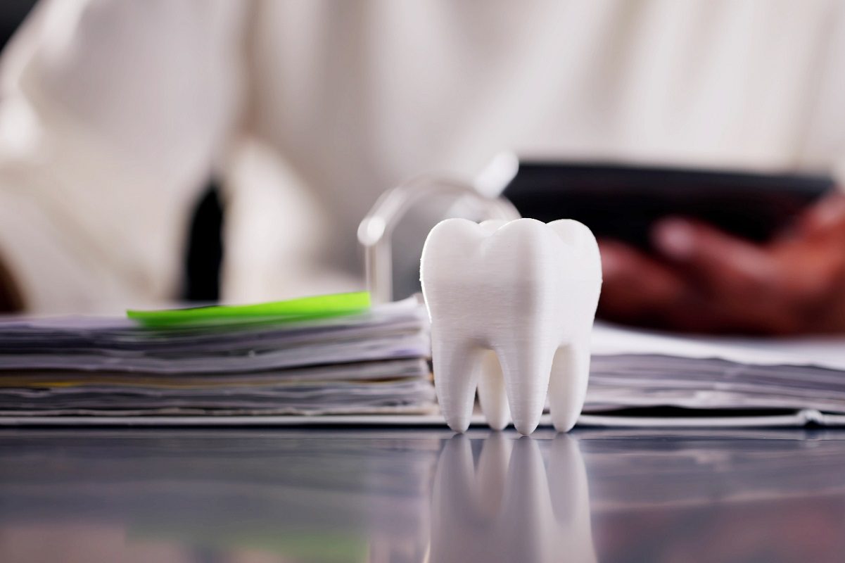 A model of a tooth stands on a reflective surface next to a stack of papers, with a person holding a tablet in the blurred background.