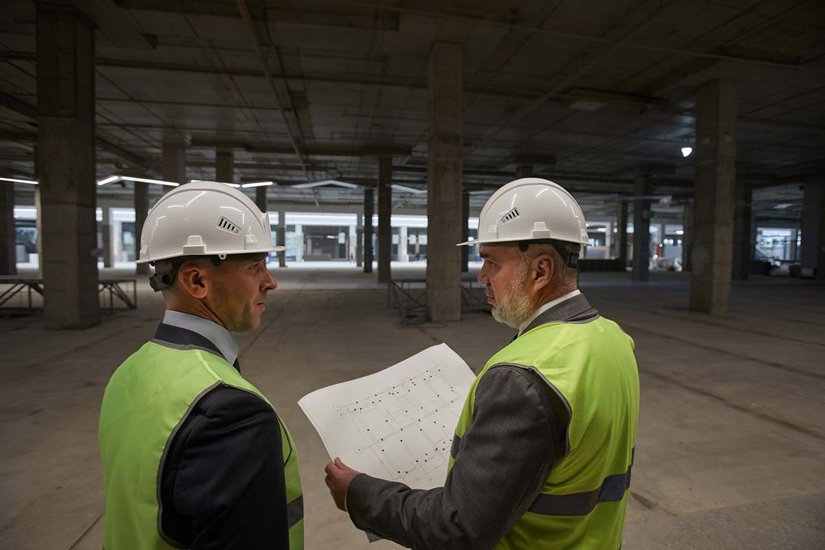 Two men in hard hats and safety vests examine a blueprint inside a large, unfinished building with concrete columns and open floor space.