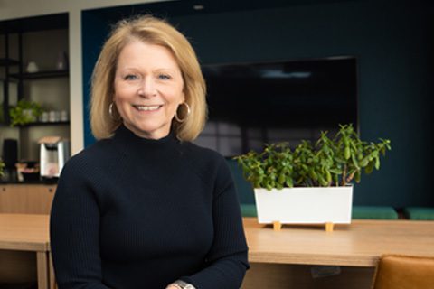 A woman with shoulder-length blonde hair wearing a black sweater sits at a wooden table with a potted plant and television in the background.
