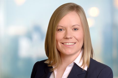 A woman with straight blonde hair wearing a dark blazer and white shirt smiles at the camera in a professional setting.