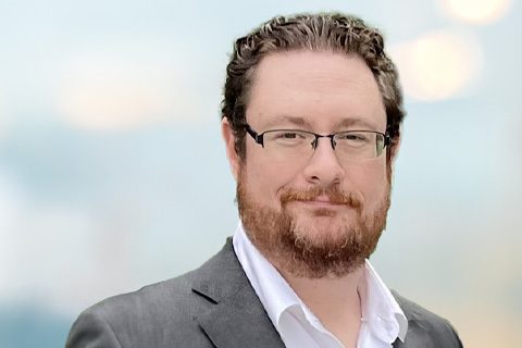 A man with short curly brown hair, glasses, a beard, and mustache, wearing a gray suit jacket over a white shirt, stands in front of a blurred light background.