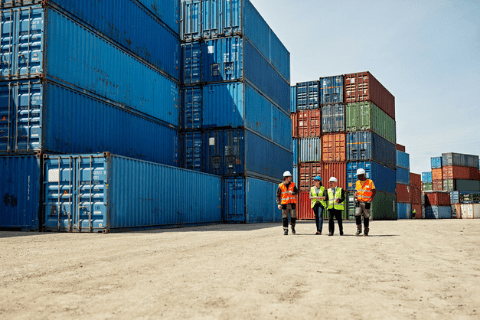 Three workers in safety gear stand near stacked shipping containers in an outdoor storage yard under a clear sky.