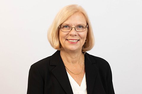 Blonde woman in glasses and a black blazer smiles in front of a white wall