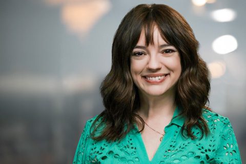 Woman with wavy brown hair wearing a green top smiles at the camera. The background is softly blurred with indistinct lights.