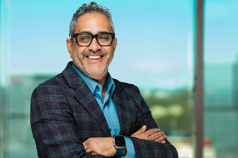 A man with gray hair and glasses is smiling, wearing a plaid blazer, blue shirt, and smartwatch, standing with arms crossed in front of a window with a blurred cityscape.