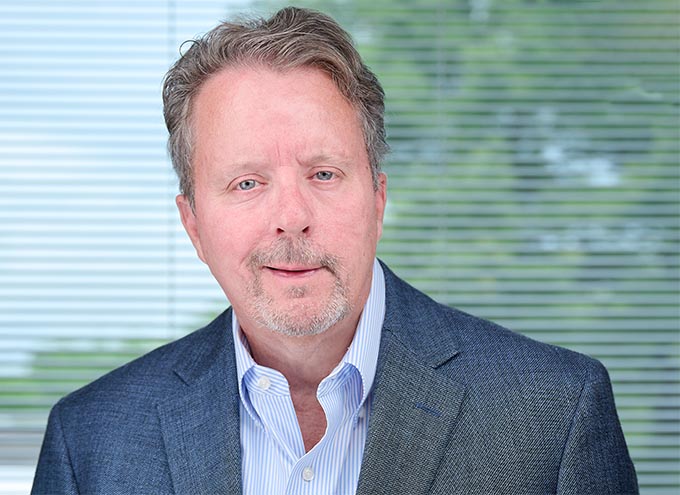A middle-aged man with short light hair and a trimmed beard, wearing a blue blazer and striped shirt, poses in front of window blinds.