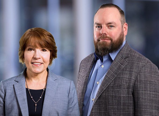 Two adults wearing business attire stand side by side in front of a blurred office background.
