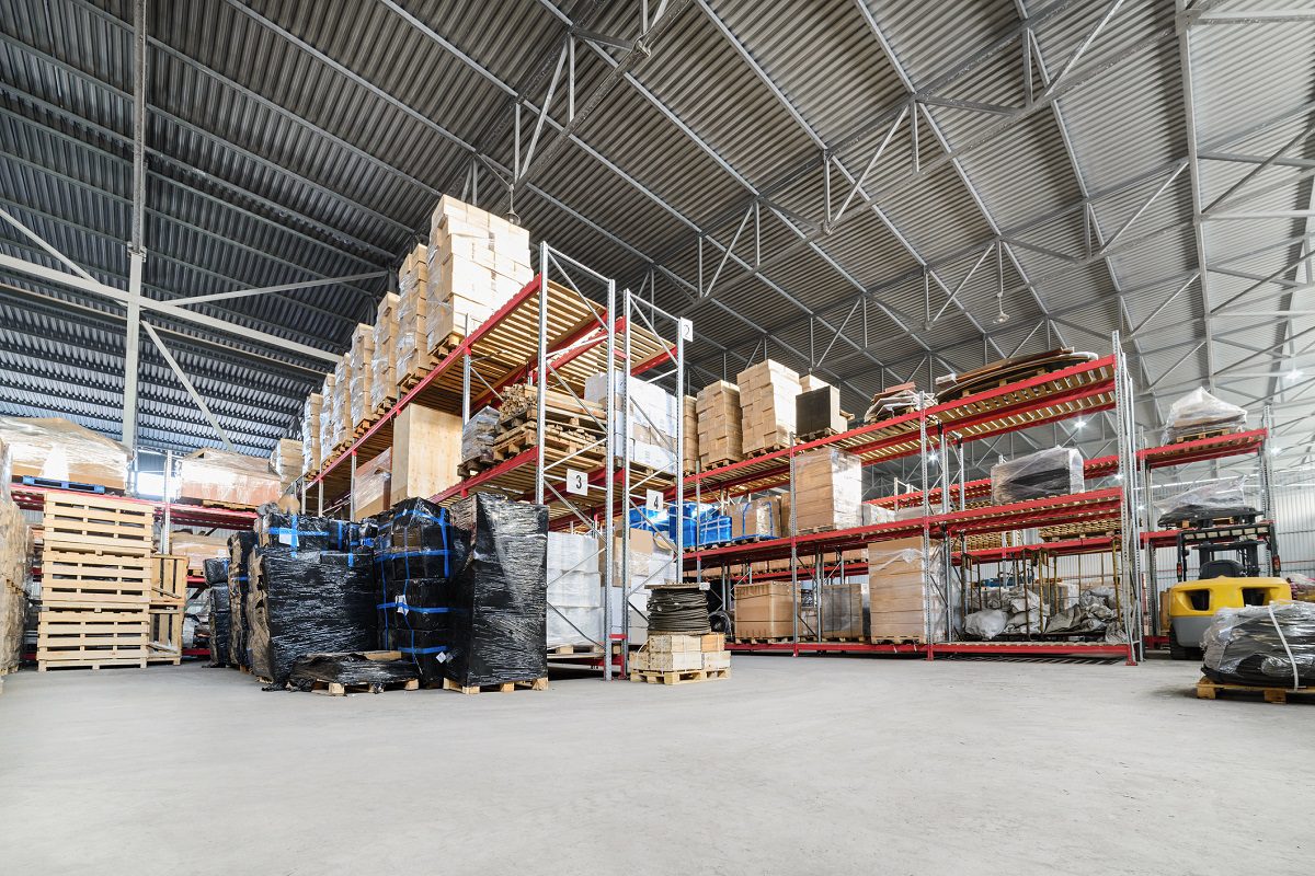 Warehouse interior with tall metal shelves holding stacked boxes, pallets, and various goods; a yellow pallet jack is visible on the right.