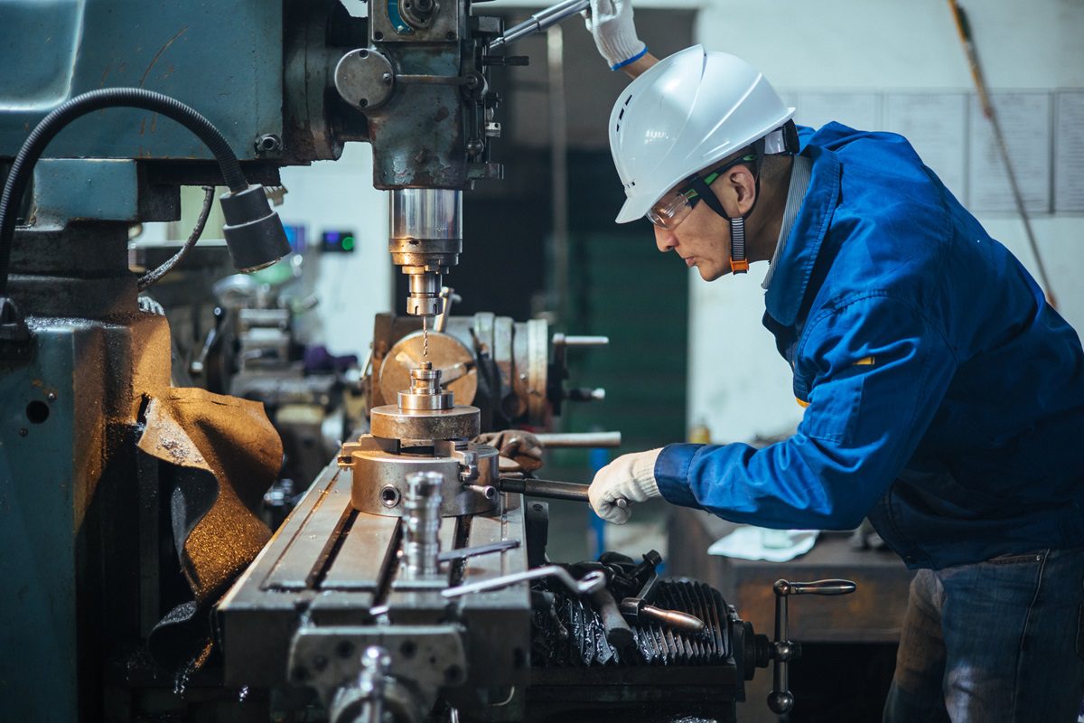 A worker in a blue jacket and white helmet operates a milling machine in an industrial workshop.