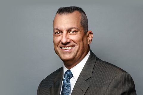A man in a dark pinstripe suit, white shirt, and patterned blue tie smiles in front of a plain gray background.
