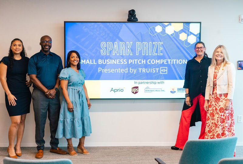 Five people stand in front of a screen displaying Spark Prize Small Business Pitch Competition Presented by Truist in a conference room.