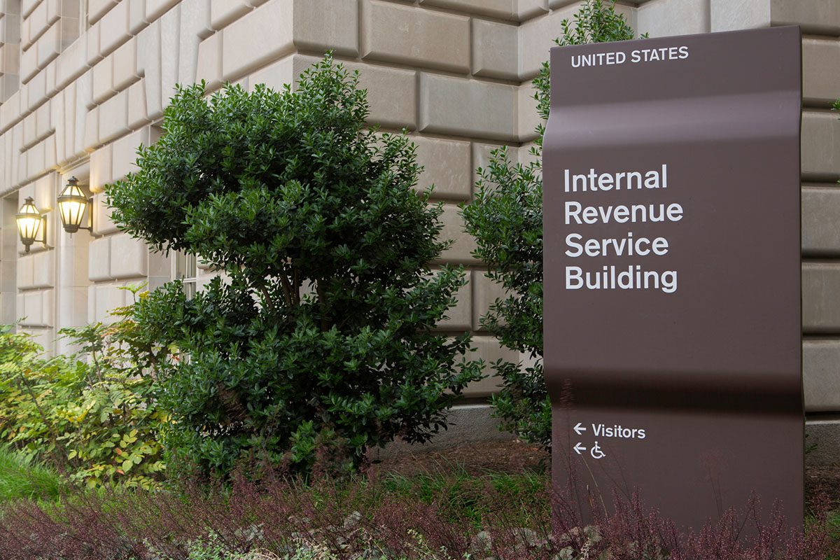 Brown sign reading Internal Revenue Service Building stands outside a stone building, surrounded by shrubs; sign includes directions for visitors and accessibility.