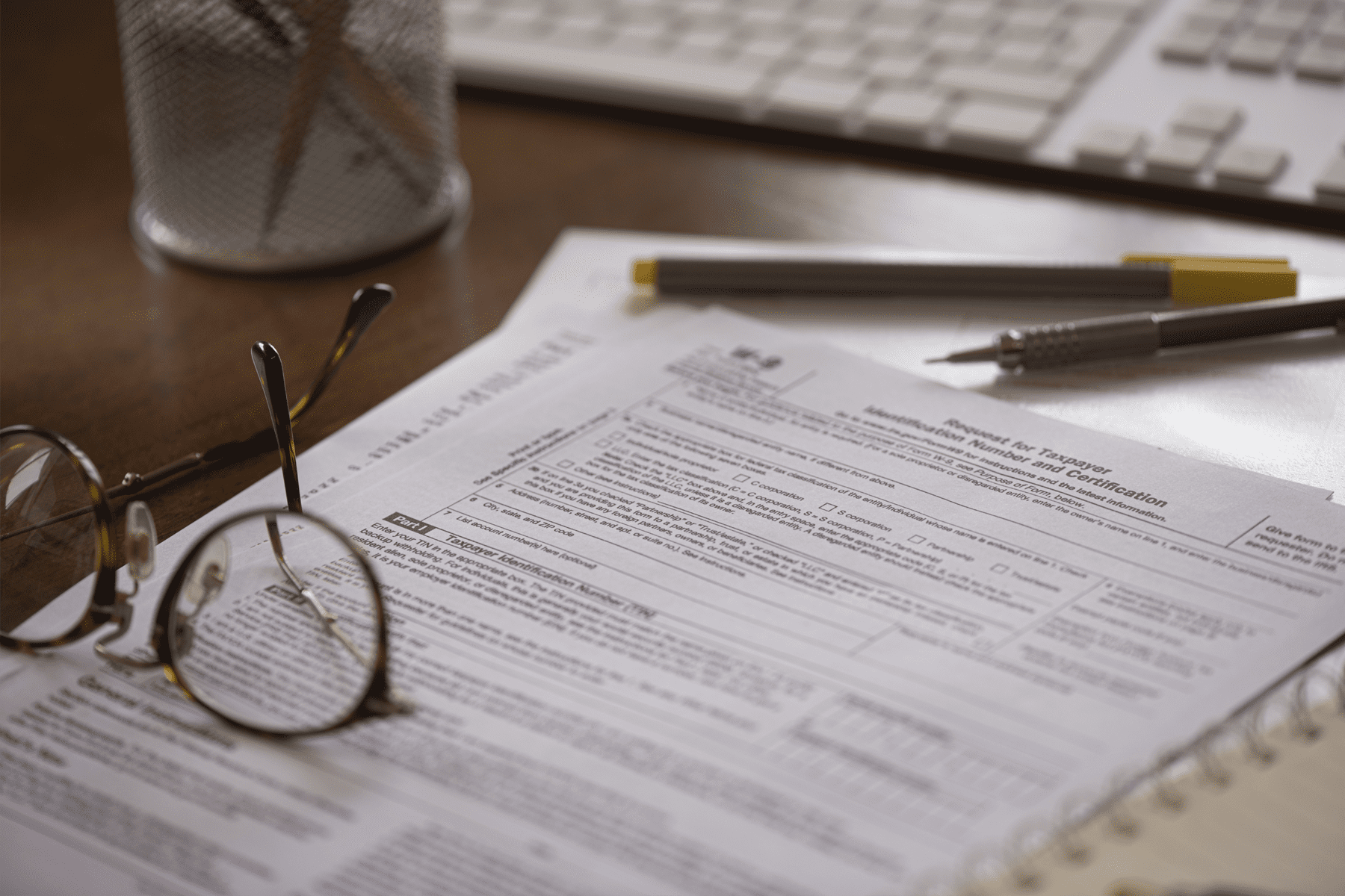 A pair of eyeglasses rests on top of tax forms, with a pen, pencil, notebook, and a keyboard in the background on a desk.