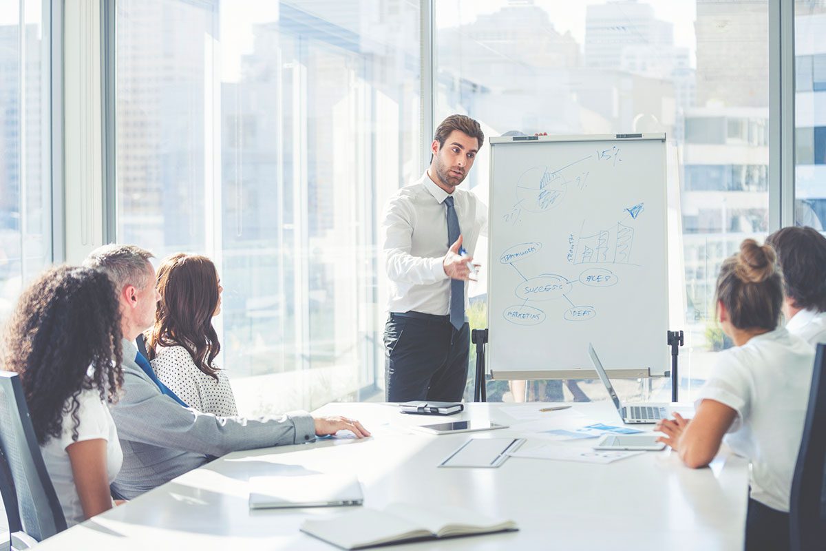A man in business attire presents a chart on a whiteboard to a group of colleagues seated around a conference table in a modern office.