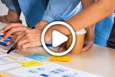 Two people work at a desk covered with notes, papers, and a coffee cup. One person writes while the other reaches across the table. A play button icon is overlaid on the image.