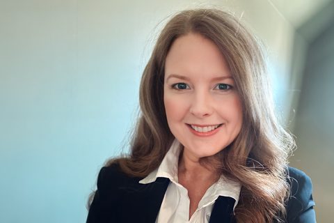A woman with long brown hair, wearing a white shirt and dark blazer, smiles at the camera against a plain light background.
