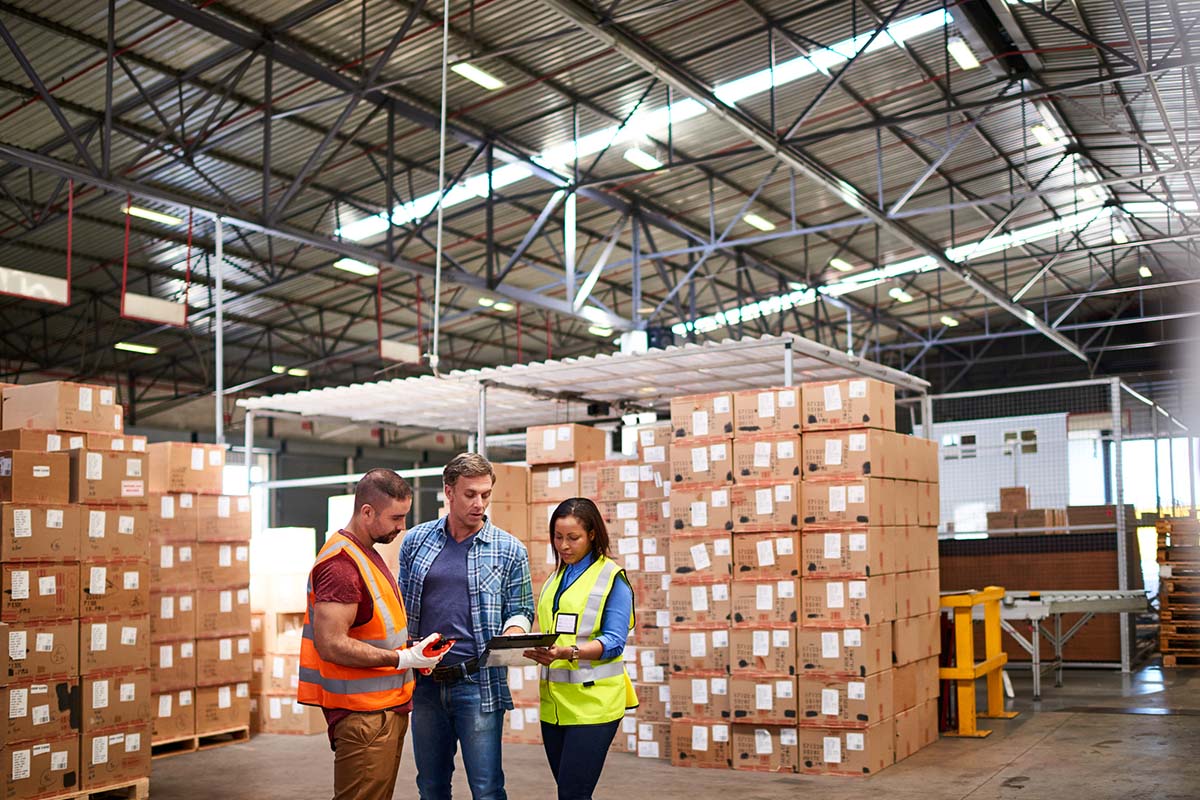 Three workers in safety vests review documents in a warehouse with stacked boxes and high ceilings.
