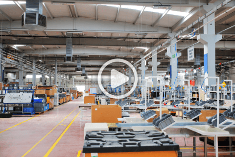 Interior of a large, modern manufacturing plant with organized workstations, industrial equipment, and overhead lighting, viewed from a central aisle.