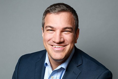 A man wearing a dark suit jacket and light blue shirt smiles at the camera against a plain gray background.