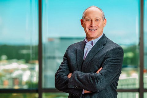 A man in a suit stands with arms crossed in front of a large window overlooking a cityscape.