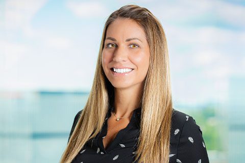 A woman with long straight hair wearing a black patterned blouse smiles at the camera against a blurred office background.