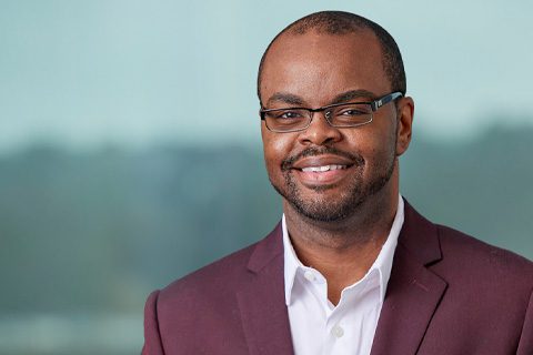 A man wearing glasses and a burgundy suit jacket over a white shirt smiles at the camera, with a blurred background.