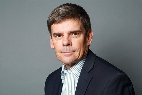 A middle-aged man with short brown hair in a dark suit jacket and striped shirt poses against a plain gray background.