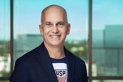 A smiling bald man in a navy blazer and a shirt with a USA soccer logo stands in front of a window with a blurred cityscape background.