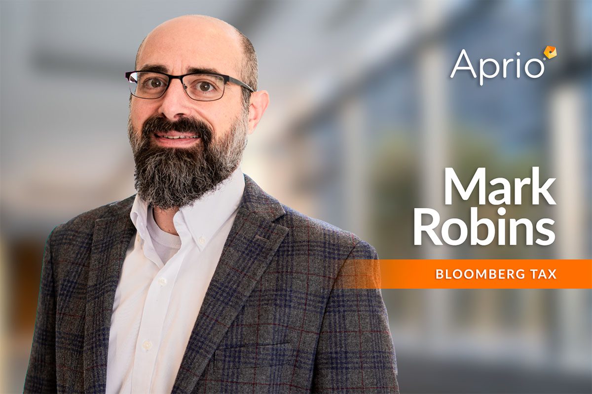 A man with glasses and a beard in a checked blazer stands in front of a blurred office background with the text Aprio, Mark Robins, Bloomberg Tax.