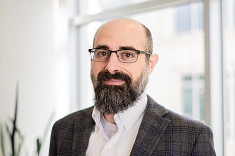 A man with glasses and a beard wearing a plaid blazer and white shirt stands indoors in front of a window.