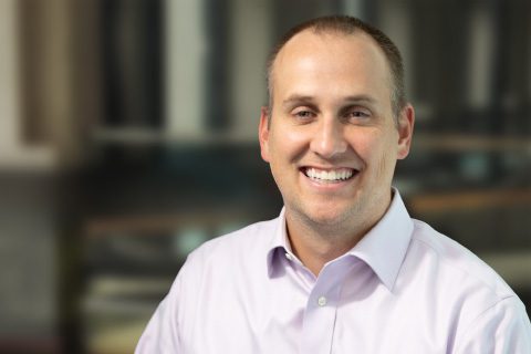 A man with short hair wearing a light purple collared shirt smiles at the camera with a blurred indoor background.