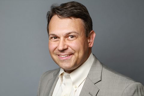 A man wearing a light gray suit jacket and white collared shirt is smiling at the camera in front of a plain gray background.
