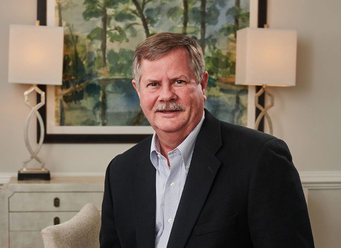 A middle-aged man with a mustache in a suit jacket and collared shirt sits indoors with lamps and a painting in the background.