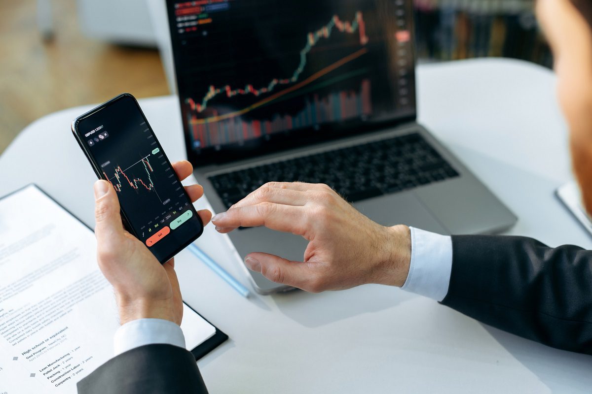 A person in a suit checks trading charts on a smartphone and laptop at a desk with documents.