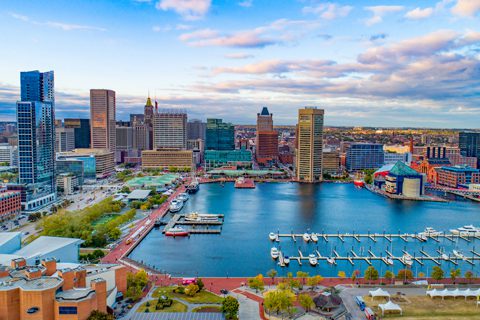 Aerial view of Baltimore Inner Harbor with waterfront buildings, docks, boats, and city skyline under a partly cloudy sky.