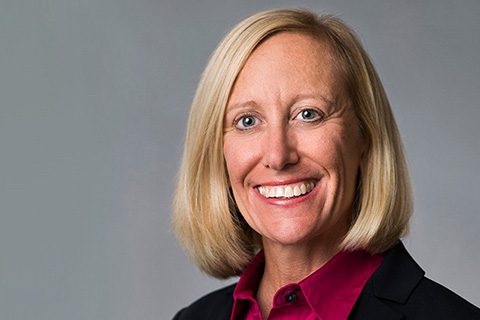 Smiling woman with straight blonde hair wearing a dark blazer and a magenta blouse, posed against a plain gray background.