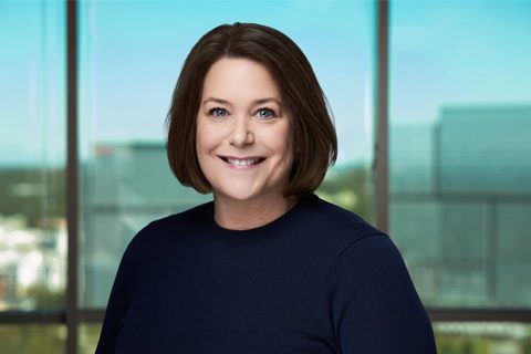 A woman with short brown hair in a navy top smiles while standing in front of large windows with an urban cityscape visible in the background.