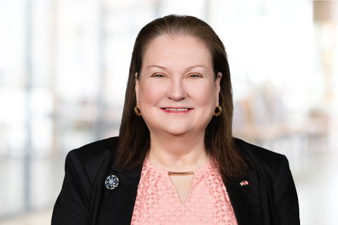 A woman with straight brown hair wearing a black blazer over a pink top, smiling at the camera with a blurred indoor background.