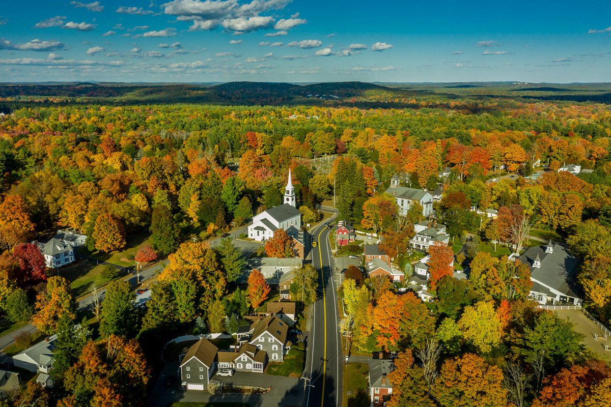 Aerial view of a small town surrounded by colorful autumn trees, with a white church and houses along a winding road under a blue sky.