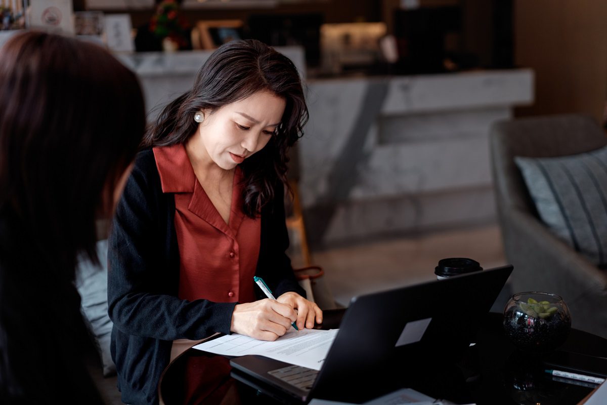 A woman sits at a table writing on documents, with a laptop and coffee cup in front of her, while another person sits nearby.