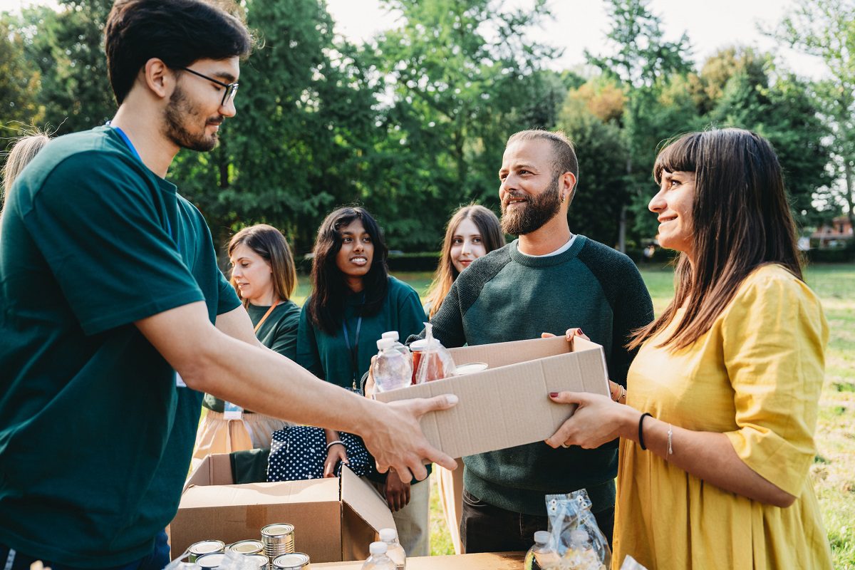 People distribute food and supplies outdoors, with one person handing a box of items to a woman while others wait nearby.