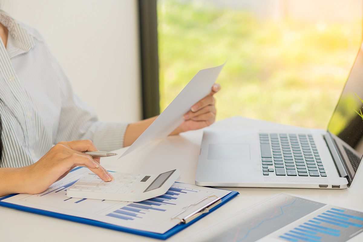 Person using a calculator with one hand and holding a document in the other, sitting at a desk with charts, a laptop, and paperwork.