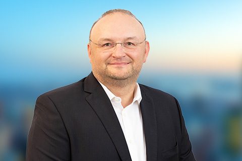 A man wearing glasses and a dark suit jacket over a white shirt is smiling at the camera, with a blurred blue and orange background.