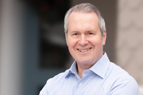 Middle-aged man with short gray hair, wearing a light blue button-up shirt, smiling and standing outdoors with blurred background.
