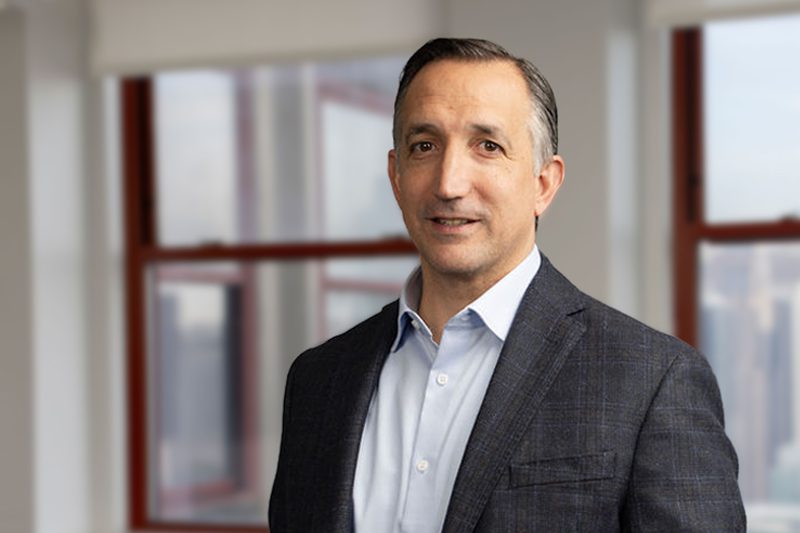 A man in a dark suit jacket and light blue shirt stands indoors in front of large windows with a city view.