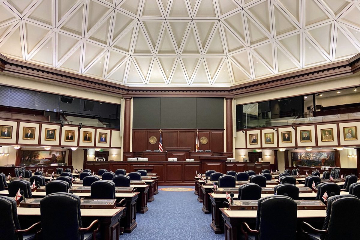 Empty legislative chamber with rows of desks and chairs, two flags behind a raised platform, framed portraits on the walls, and a geometric-patterned ceiling.