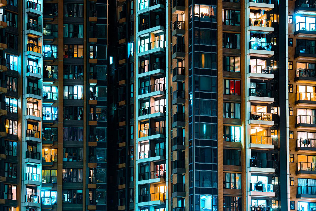 High-rise apartment building at night with numerous illuminated windows in various colors, showing glimpses of different rooms and balconies.