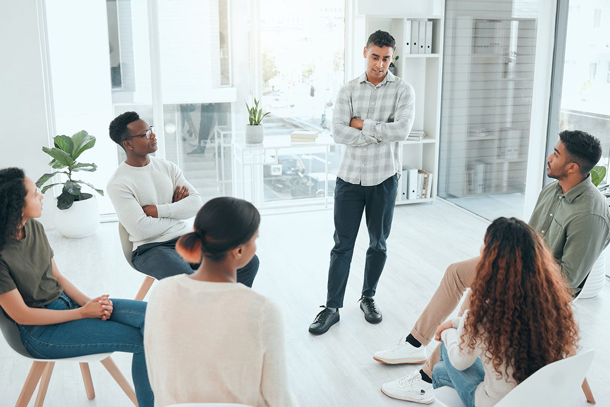 A man stands and speaks to five seated people in a bright, modern office, suggesting a group discussion or meeting.