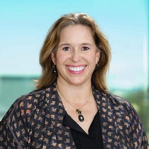 A woman with shoulder-length light brown hair, wearing a patterned jacket, black top, and a necklace, smiles at the camera against a blurred indoor background with a window.