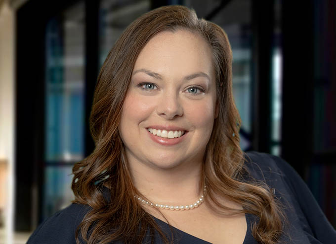 Woman with long brown hair, wearing a navy blue top and a pearl necklace, smiling in front of a blurred indoor background.